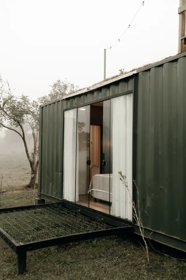 Casa hecha con un contenedor verde, con puertas de vidrio, en un campo con niebla